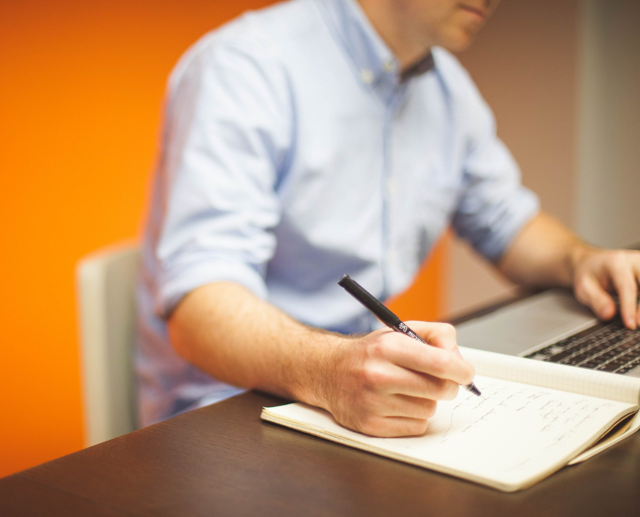 Man in an office writing noted in a book while looking at his laptop